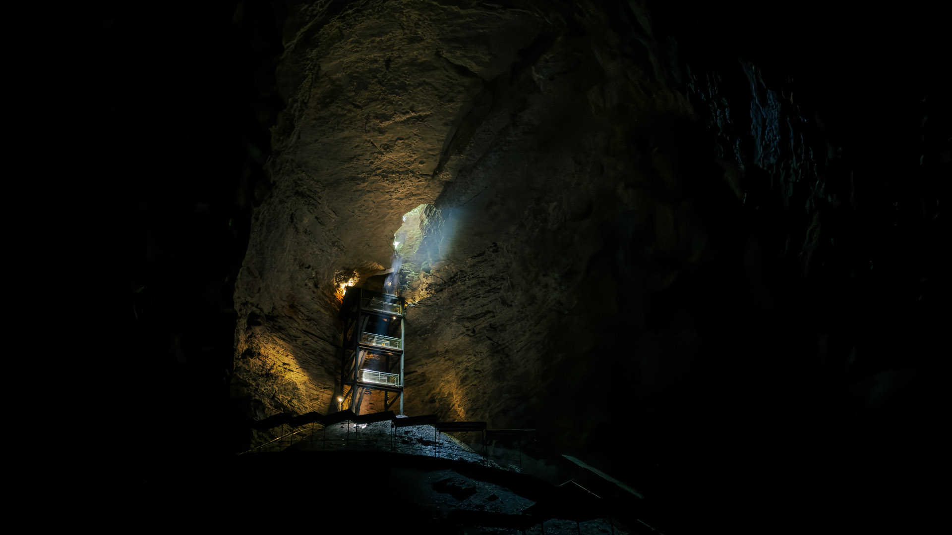 Dark cave interior with a lit stair tower and daylight entering from above